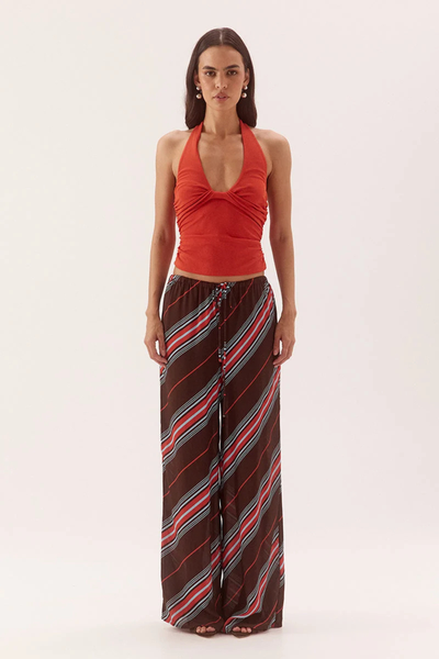 Full-length studio front view of a model wearing a bright vermilion red halter neck top featuring front bust tucks and waist gathers, styled with wide-leg dark brown striped trousers.