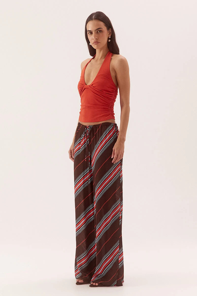 Studio three-quarter front view of a model wearing a vermilion red halter top with a deep scoop neckline and side bodice gathers, paired with wide-leg brown striped trousers.