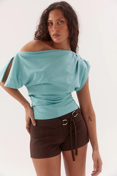 Studio medium shot of a model wearing a glacier blue pure cotton asymmetric tee, draped off one shoulder with a split sleeve detail, paired with dark brown shorts.