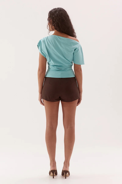 Studio full-length rear view of a model wearing a glacier blue cotton tee showcasing the draped off-shoulder neckline, waist gathering, and short sleeve structure.