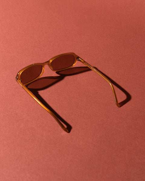Top-down perspective studio shot of the Sito Shades Roux sunglasses in translucent caramel orange acetate displaying temple arms and lens shadow against a terracotta background.