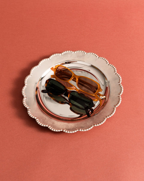 Overhead studio flat-lay of the Sito Shades Roux sunglasses in translucent orange caramel acetate alongside a dark tortoiseshell pair on a scalloped silver serving tray over a terracotta pink surface.
