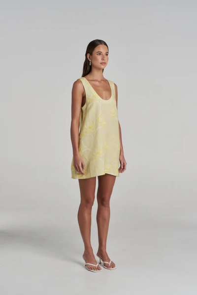 Full-length studio side profile view of a model wearing the yellow linen-blend Flora Mini Dress with a white floral print and a scoop neckline, paired with heeled sandals.