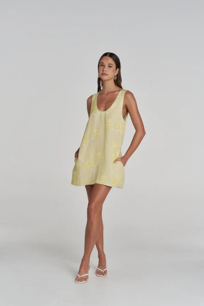 Full-length studio front view of a model posing with her hands in the pockets of the yellow floral mini dress, paired with white heeled sandals.