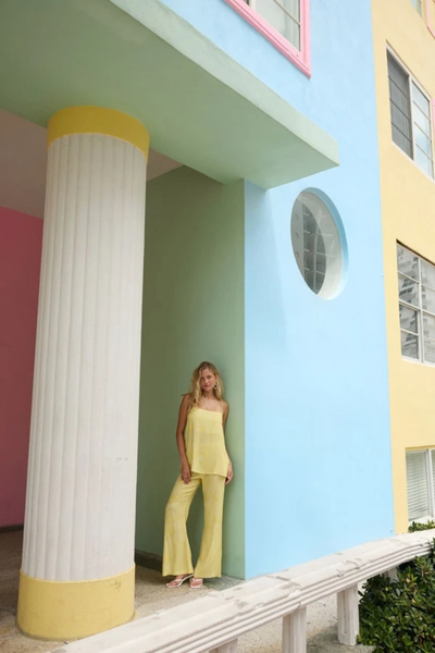 Full-length lifestyle shot of a model leaning against a pillar in a colorful pastel building, wearing the matching yellow floral camisole and flare pants set with white sandals.
