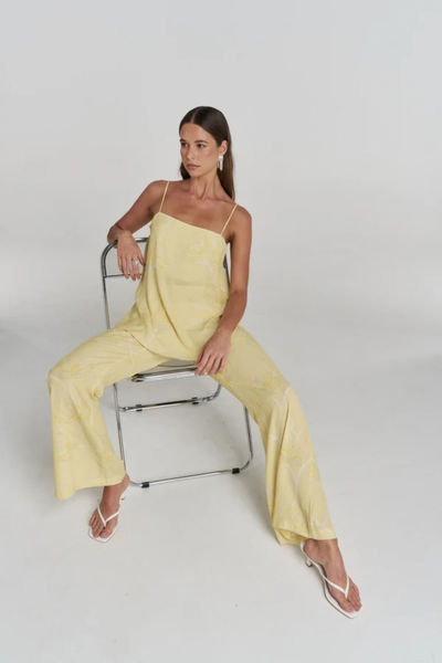 Studio view of a model sitting on a chrome chair, wearing the matching yellow floral set and white heeled sandals.