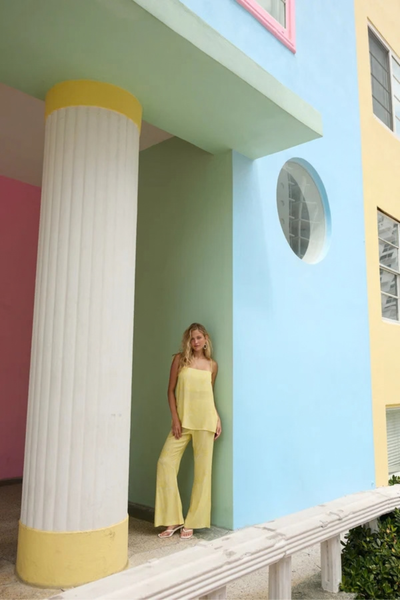Full-length lifestyle shot of a model leaning against a pillar of a colorful pastel building, wearing the matching yellow floral tunic and pant set.