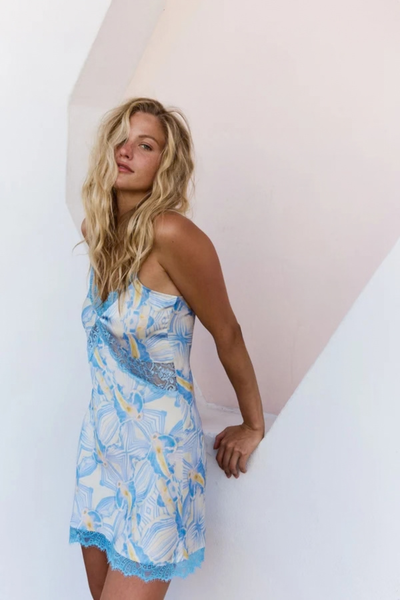 Close-up lifestyle portrait of a model with blonde wavy hair leaning against a modern architectural wall, wearing the blue and white patterned slip dress with blue lace trim.
