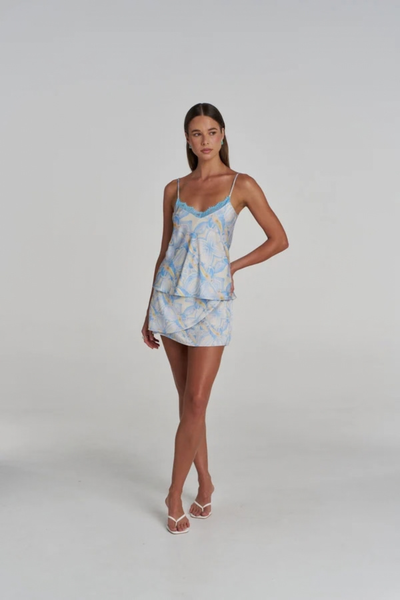 Full-length studio front view of a model standing straight, wearing the matching blue and white Orchid Prism lace camisole and mini skirt set with white heeled sandals.