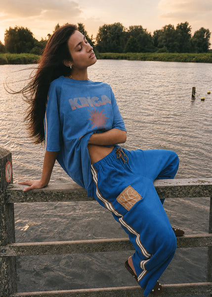 Outdoor lifestyle shot of a model wearing the Nuwa Blue Sunset Tee and matching slouchy blue track pants with a tan "KC" patch. She is lounging on a rustic wooden railing by a lake at sunset, showing the sporty, lived-in aesthetic of the set.