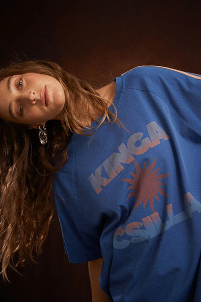 Front-on studio portrait of a model wearing the relaxed Nuwa Blue Sunset Tee. The shot highlights the loose-fit silhouette, the crew neck, and the contrasting tan stripes running down the shoulders.
