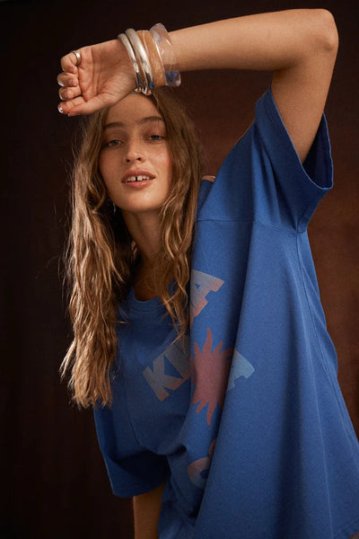 Studio portrait of a model with wavy blonde hair wearing the relaxed Nuwa Blue cotton Sunset Tee against a dark brown background. She poses with her arm raised over her forehead, highlighting the comfortable drape of the raglan sleeve and chunky silver bangles.