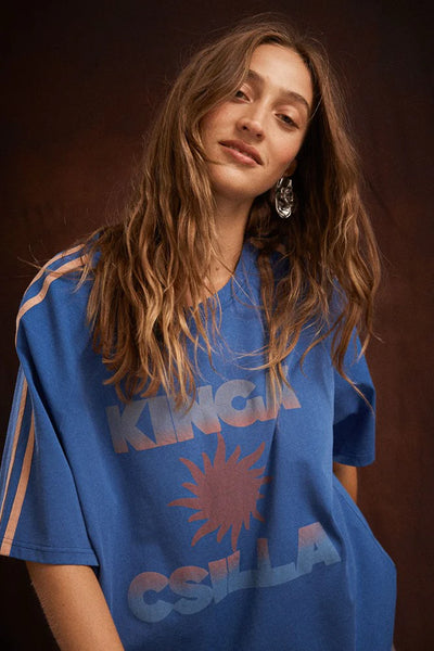 Dynamic studio shot of a model wearing the relaxed-fit Kinga Csilla Sunset Tee in Nuwa Blue. Leaning slightly to the side against a dark, moody backdrop, she showcases the casual crew neckline and the faded sunburst gradient graphic.