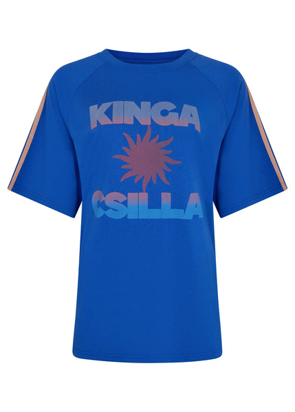 Studio e-commerce product shot of the Kinga Csilla Sunset Tee in Nuwa Blue against a clean white background. This front view showcases the classic raglan sleeve cut, crew neckline, tan shoulder stripes, and the central coral-to-sky-blue gradient sunburst graphic.
