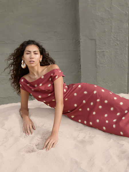 An editorial campaign shot of a model with curly hair reclining on a sand dune against a textured gray wall, showcasing the effortless drape of the brick red, polka-dot Saro dress.