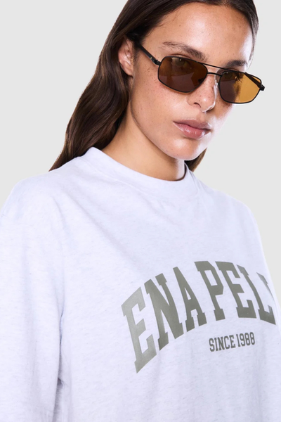 Close-up portrait of a model wearing sunglasses and a white marle crew-neck t-shirt highlighting the olive green arched "Ena Pelly" chest text print.