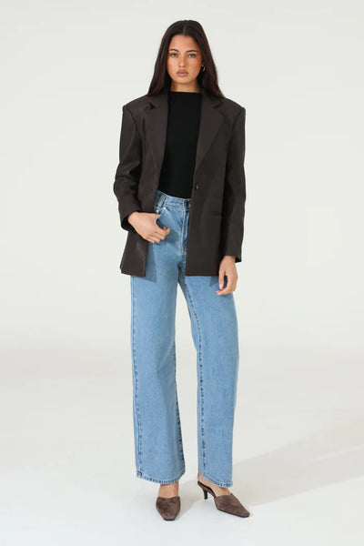 A full-length studio portrait of a model presenting the chocolate brown PU leather Sutton Blazer buttoned at the center front, showcasing its relaxed silhouette, exaggerated collar, and sharp lapels styled with a black high-neck top, light-wash wide-leg jeans, and brown pointed-toe mules.