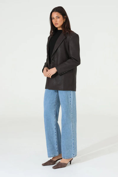 A full-length rear three-quarter studio view of the model wearing the oversized chocolate brown faux leather blazer, emphasizing its fluid, structured drape, horizontal back-seam detail, and button-cuffed long sleeves.