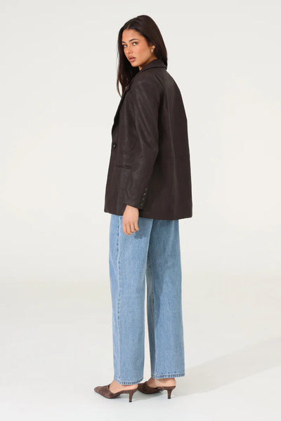 A full-length angled studio shot of the model wearing the rich chocolate PU leather tailored jacket, highlighting its comfortable relaxed fit, pocket flap details, and sophisticated look from a side profile.