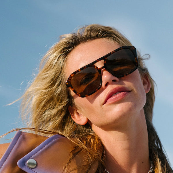 A close-up lifestyle campaign photograph of a blonde model captured outdoors against a clear blue sky. She is looking downward at the camera with a slight head tilt, wearing the tortoise shell aviator sunglasses paired with a leather-look collared jacket in shades of tan and lavender, projecting a sun-drenched vintage aesthetic.