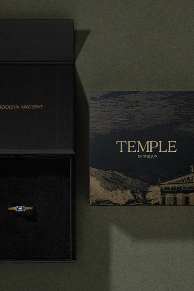Flat-lay packaging shot of the Temple of the Sun jewelry box and branded gift card on a textured green background.