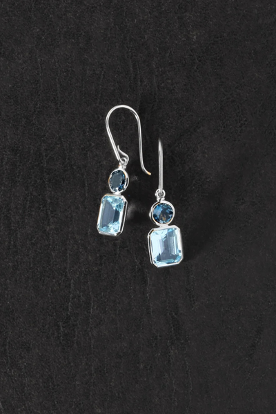 Overhead flat-lay shot of the pair of Temple of the Sun Aqua Earrings in Sterling Silver displaying the blue topaz stones and silver hook details on a textured dark background.