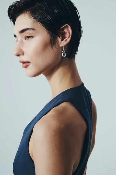 Side-profile portrait of a short-haired model wearing the Temple of the Sun Sterling Silver Aqua Drop Earrings paired with a dark blue top.