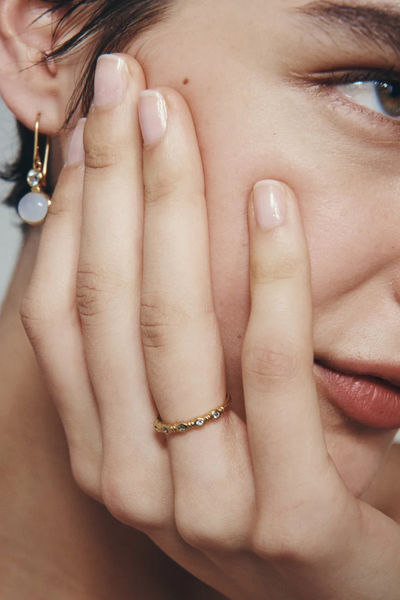 Close-up profile of a model touching her face, wearing the Temple of the Sun Aqua Ring in 18k Gold Vermeil on her finger alongside matching gold drop earrings.