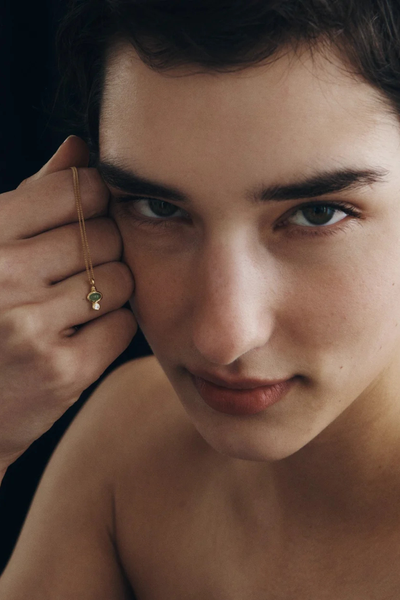 Close-up shot of a model holding the delicate pendant of the Temple of the Sun Ma Necklace in 18k Gold Vermeil next to her face.