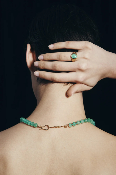 Lifestyle portrait showing a model wearing the Temple of the Sun Ma Ring in 18k Gold Vermeil on her hand rested behind her head, paired with a green gemstone necklace.