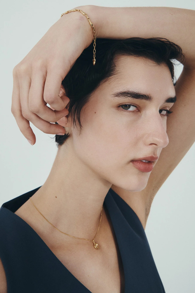 Lifestyle shot of a model wearing the Soma Coin Necklace in 18k Gold Vermeil with her hand rested over her head, showcasing a gold link bracelet.