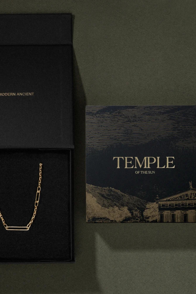 Top-down view of an open black Temple of the Sun branded gift box displaying a chain beside an artwork card on a green background.
