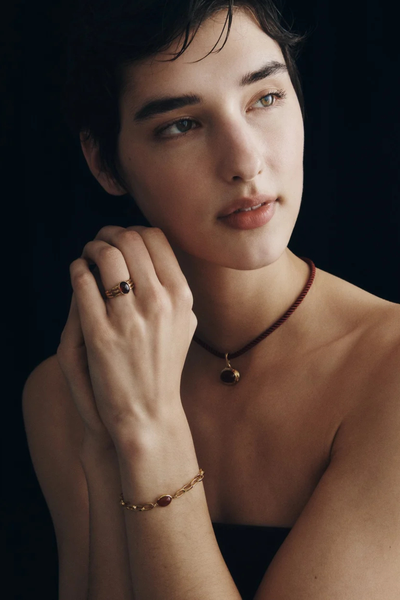Model posing with hands held together, wearing the Vega Garnet Ring, a matching chain bracelet, and a cord necklace.