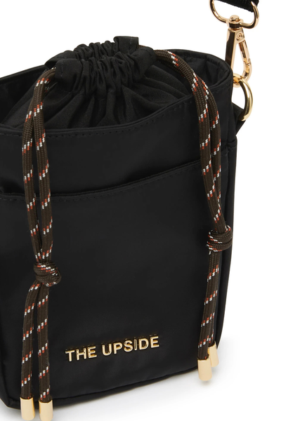 Close-up studio detail shot of a black recycled polyester crossbody bag featuring an internal drawstring closure with chocolate paracord cords, a front slip pocket, gold metal hardware, and a gold metal "THE UPSIDE" logo decal.