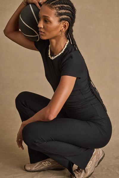 Side profile view of a model crouching while holding a black basketball, wearing the black flare pants with hem splits, a black t-shirt, and sneakers.