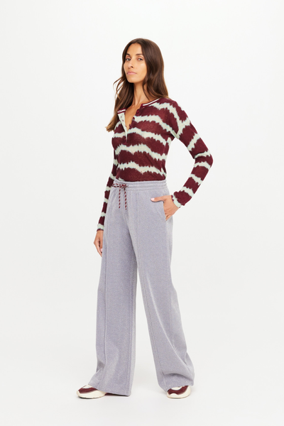 Full-length campaign shot of a model wearing the Galaxia Marnie Wide Leg Pants. The shimmering grey pants are styled with a white rib-knit tank top, a draped tie-dye pullover, and layered gold chain necklaces for an effortless, street-chic athleisure look.