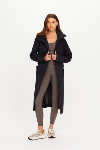 Full-length front view of a model showcasing the open silhouette of The Upside James Hooded Trench Coat in black sand. The longline, weather-resistant recycled polyester trench is worn open over a brown and black houndstooth print jumpsuit and neutral athletic sneakers, highlighting its relaxed, layerable design.