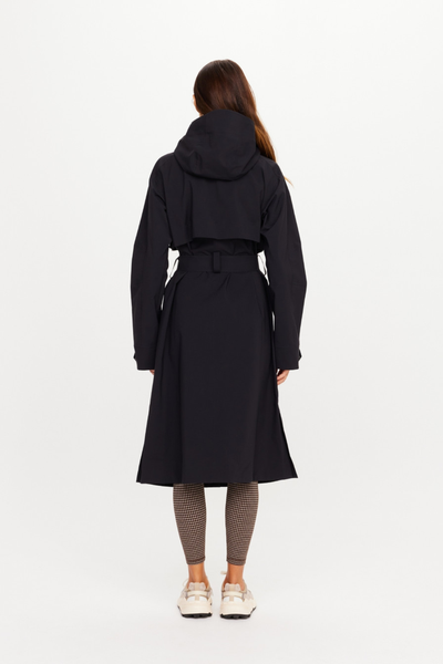Full-length rear view of a model wearing the closed, belted James Hooded Trench Coat in black sand. The shot highlights the structured back storm flap yoke, the relaxed hang of the hood, and the elegant, functional longline drape of the coat.