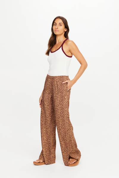 Full-length studio shot of a model wearing the Javan Juliet Pant in Javan leopard print, paired with a white ribbed tank top featuring burgundy trim. The lightweight, wide-leg twill pants feature a relaxed fit and functional side seam pockets.