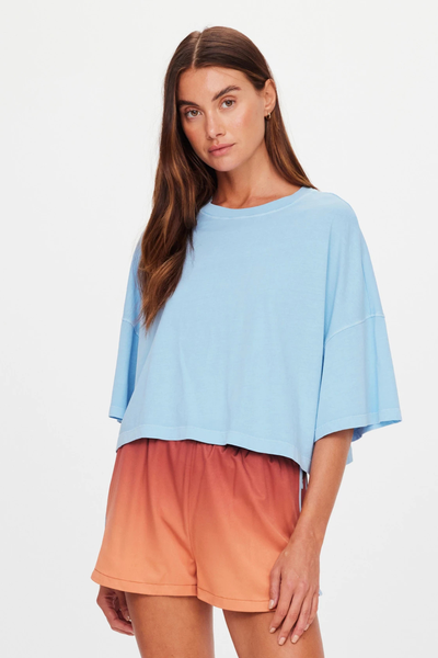 Studio three-quarter front view of a brunette model wearing a light blue overdyed organic cotton cropped boxy t-shirt styled with red-to-orange ombre running shorts.