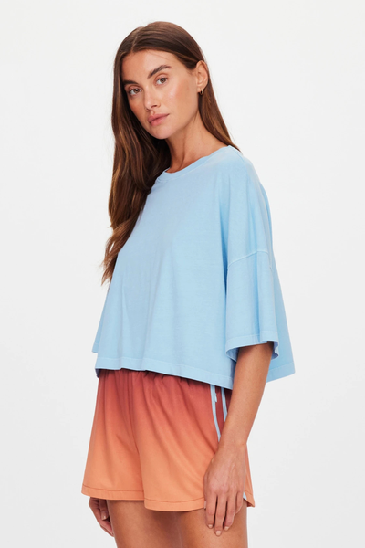 Studio side profile portrait of a model wearing an overdyed light blue cropped t-shirt featuring wide elbow-length drop-shoulder sleeves.
