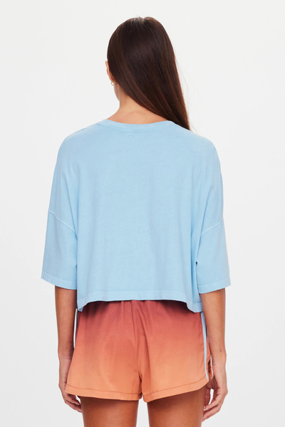 Studio rear view of a model wearing a boxy overdyed light blue organic cotton cropped t-shirt paired with sunset ombre shorts.