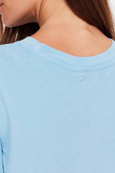 Close-up studio rear detail shot highlighting the ribbed neckline and subtle tonal embroidered arrow logo on the back upper neck of a light blue cropped tee.