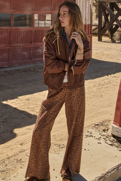 Full-length outdoor campaign image of a model wearing the chocolate brown Lila Zip Through Jacket. She is styled in a desert setting with matching leopard-print wide-leg pants and a white tank top, highlighting the jacket's casual, street-ready styling.