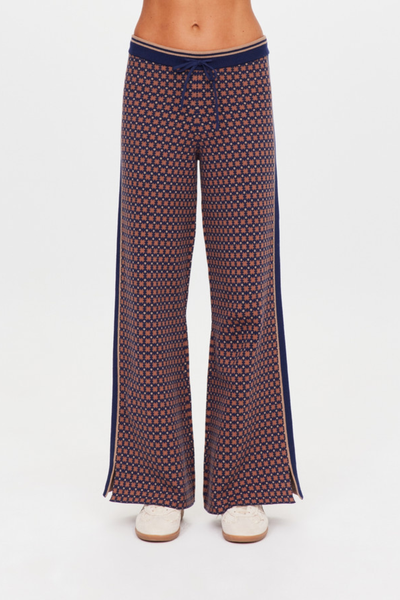 A mid-section studio close-up focusing on the waistband and hip of the Lumiere Juniper Knit Pant. The image showcases the organic cotton blend knit jacquard fabric in a navy blue and tan geometric star design, emphasizing the ribbed navy waistband with contrast tan stripes and a matching navy drawcord tie.
