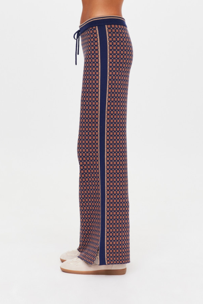 A side-profile studio shot isolating the Lumiere Juniper Knit Pant to showcase the continuous, solid navy knit panel running down the side seam, flanked by thin tan contrast stripes, as well as the structural side splits at the bottom hem.