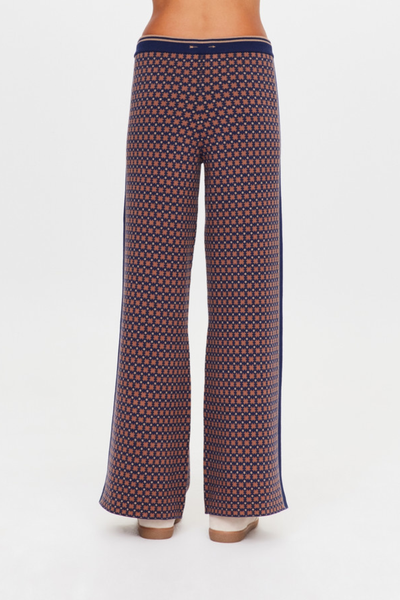 A full-length studio shot from the back showing a model wearing the high-waisted geometric star pattern knit trousers. The photo highlights the straight-leg drape down to the ankles, the clean rear profile, and the signature tonal embroidered arrow logo positioned centered on the back waistband.