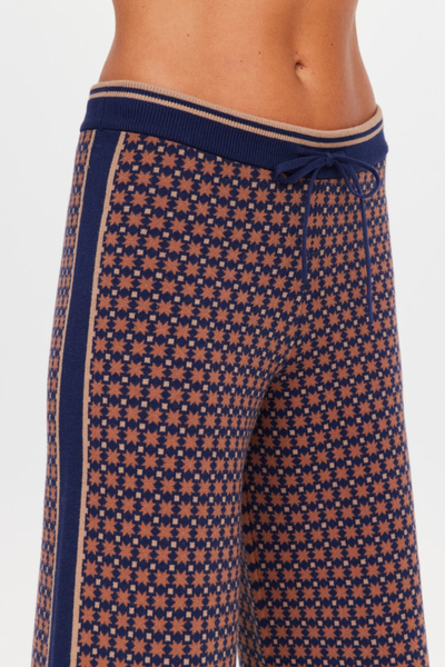 A mid-section studio close-up shot focusing on the high-waisted fit and geometric star jacquard pattern of the Lumiere Juniper Knit Pant. The image details the flexible, ribbed navy blue waistband with its contrasting tan trim, the functional matching drawstring tie, and the solid navy side-stripe panel flanked by thin tan accent lines down the outer leg seam.