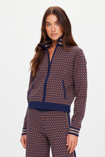 A three-quarter studio close-up shot focusing on the design details of the Lumiere Margot Knit Jacket. The image highlights a model with her hand in one of the front welt pockets, showcasing the organic cotton blend knit jacquard fabric in a navy blue and tan geometric star pattern, along with the ribbed navy waistband and matching striped cuffs.
