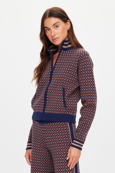 A three-quarter profile studio photograph of a model wearing the Lumiere Margot Knit Jacket in Geo, styled zipped up with a high mock-neck collar. The shot highlights the relaxed silhouette, structured front pockets, and the sporty, striped elastication along the sleeve cuffs and waistband.